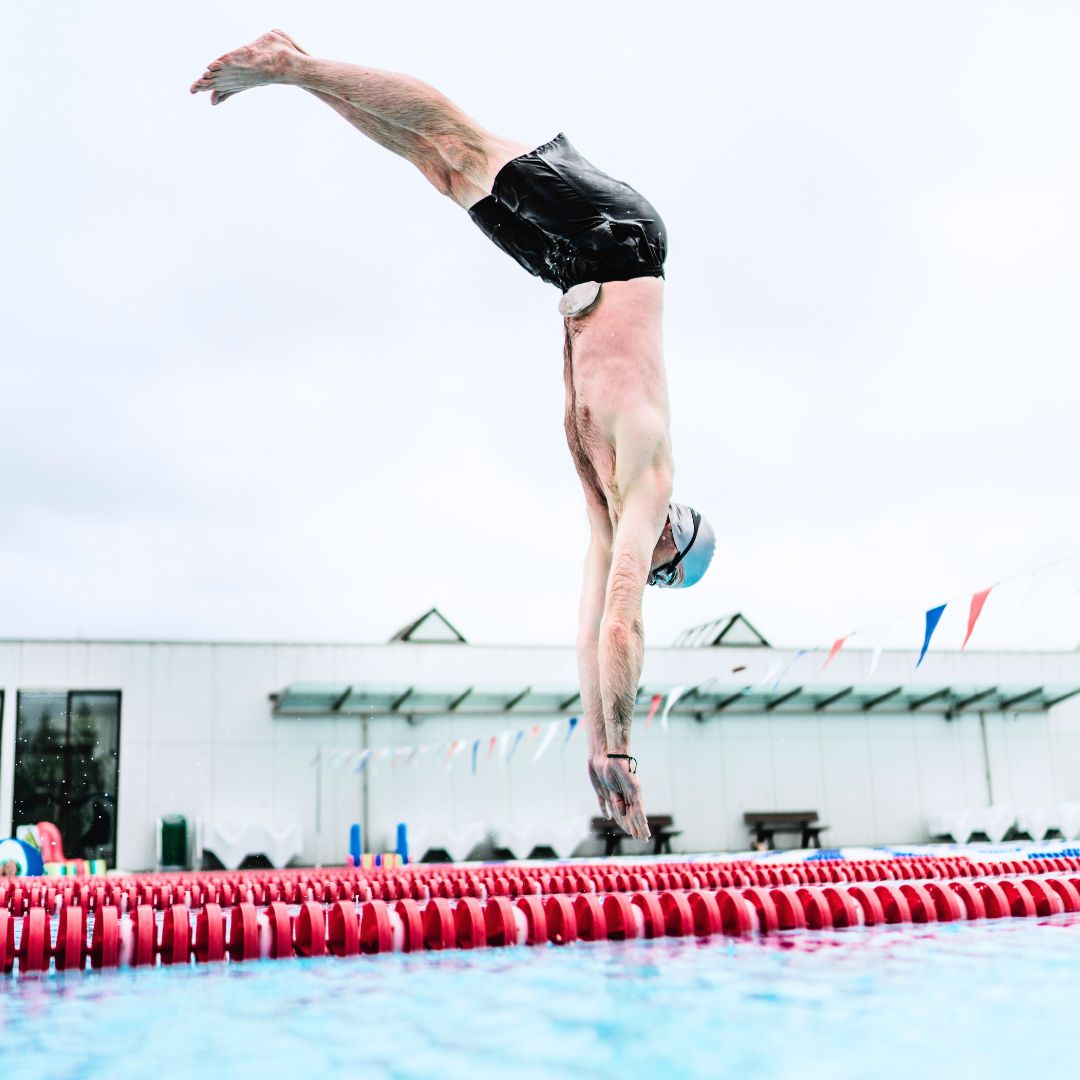 Osoba wskakująca do basenu w stroju kąpielowym, uchwycona w ruchu nad taflą wody w słoneczny dzień.